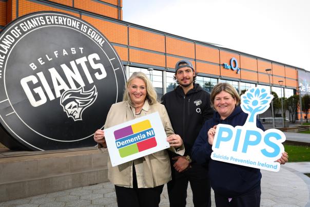 Three people stand holding charity signs in front of a Belfast Giants sign and a large orange building.
