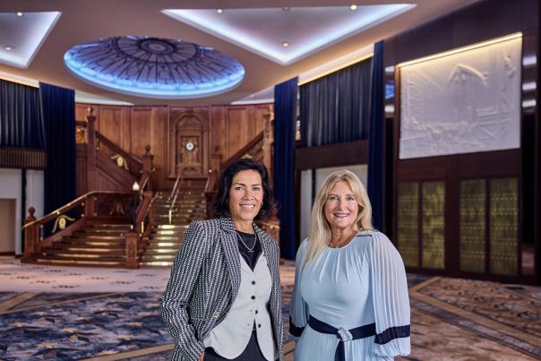 Two women stand in a grand foyer with a sweeping staircase and ornate ceiling in the background.