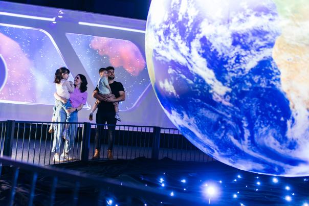 A family stands on a walkway in the foreground, looking at a large, illuminated globe of Earth in the background, with a starry, cosmic display on a screen behind it.