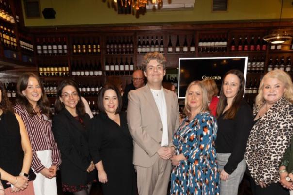 A group of eight people, smiling, stand closely in a warmly lit room with shelves of wine bottles.