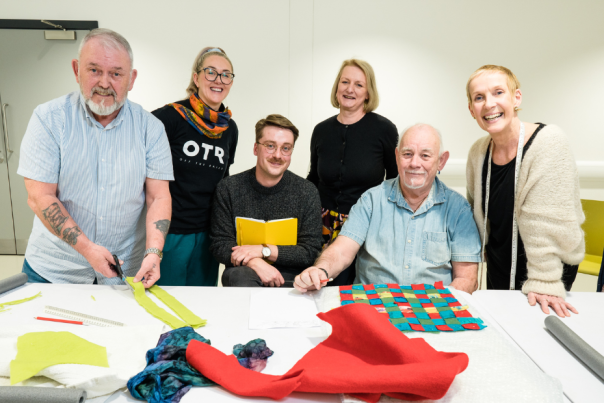Six people smile for a photo behind a white table covered in colorful fabric scraps and sewing tools against a plain white wall.