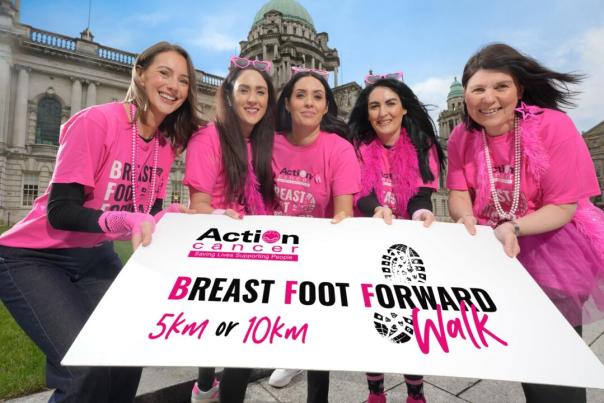 A group of smiling women wearing matching bright pink shirts pose together outdoors, holding a large sign that reads “Action Cancer Breast Foot Forward Walk 5K or 10K."
