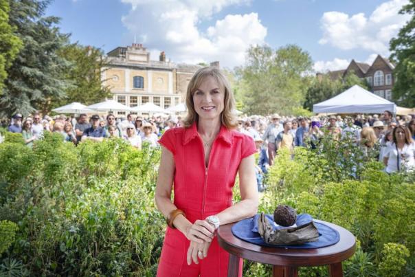 Woman stands by an antique on a table, with a crowd behind her at an Antiques Roadshow event.