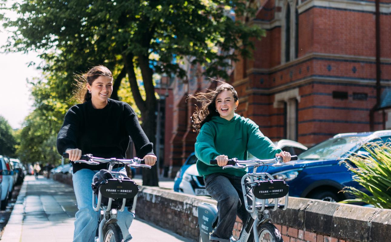 Two female students cycling past Queen's University on Belfast Bikes