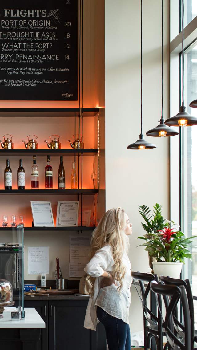 Blonde haired woman staring out the window of a coffee shop with a counter filled with pastries.  There is a chalkboard menu and wine bottles on the wall, and stylish pendant lights hanging from the ceiling