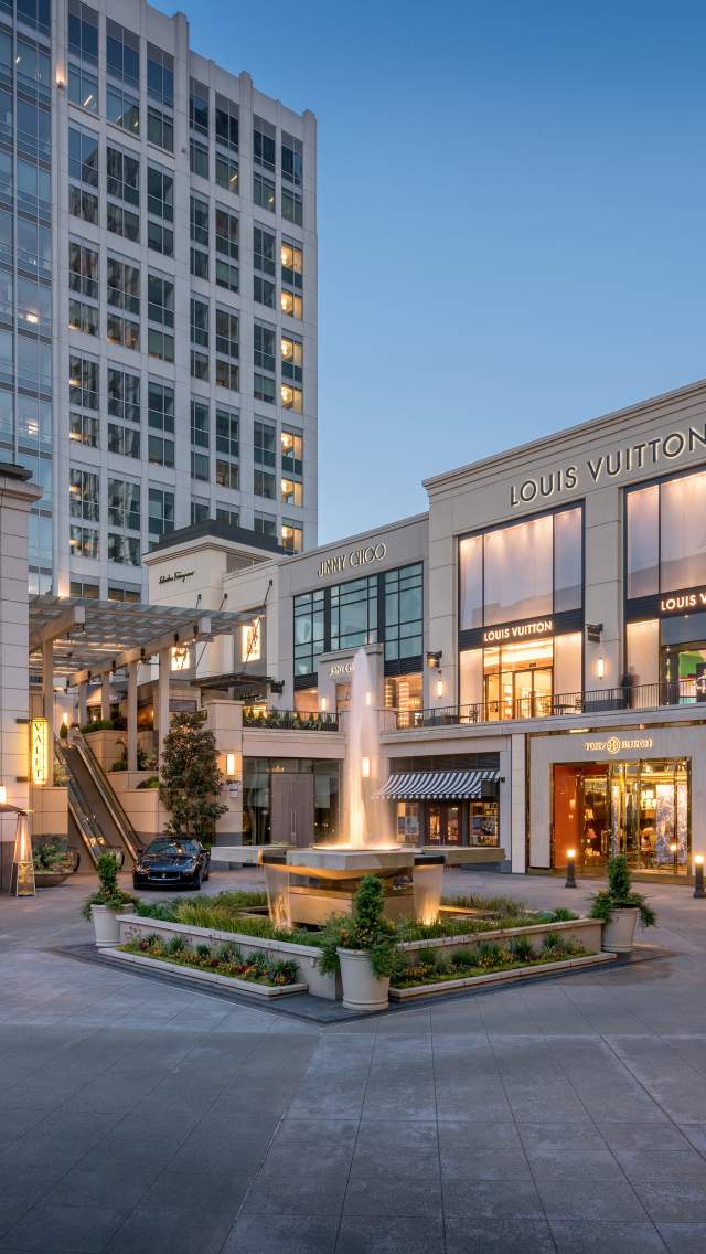 Bravern Bellevue luxury shopping plaza featuring Hermes, Louis Vuitton, Prada, with a fountain and modern architecture at dusk.