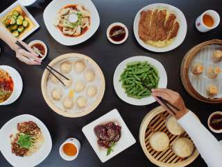 Black wood table full of brightly colored food, and dumplings in wooden baskets, hands reaching across the table with chopsticks