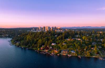 Aerial view of a lush, tree-covered peninsula in Bellevue with houses along a calm lake at sunset, the city skyline in the background beneath a pink and purple sky, creating a serene atmosphere.