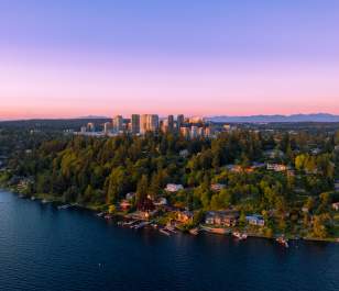 Aerial view of a lush, tree-covered peninsula in Bellevue with houses along a calm lake at sunset, the city skyline in the background beneath a pink and purple sky, creating a serene atmosphere.