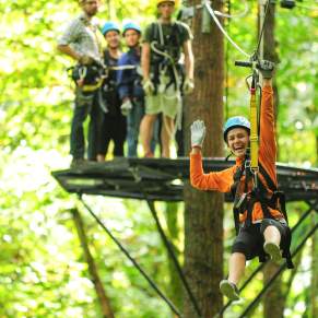 A person in orange zip-line gear is gliding through a lush green forest, with others waiting on a platform in the background.