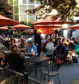 A lively Bellevue outdoor dining area filled with patrons seated under orange umbrellas, enjoying food and drinks in a relaxed atmosphere.