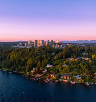 Aerial view of a lush, tree-covered peninsula in Bellevue with houses along a calm lake at sunset, the city skyline in the background beneath a pink and purple sky, creating a serene atmosphere.