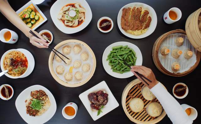 Black wood table full of brightly colored food, and dumplings in wooden baskets, hands reaching across the table with chopsticks