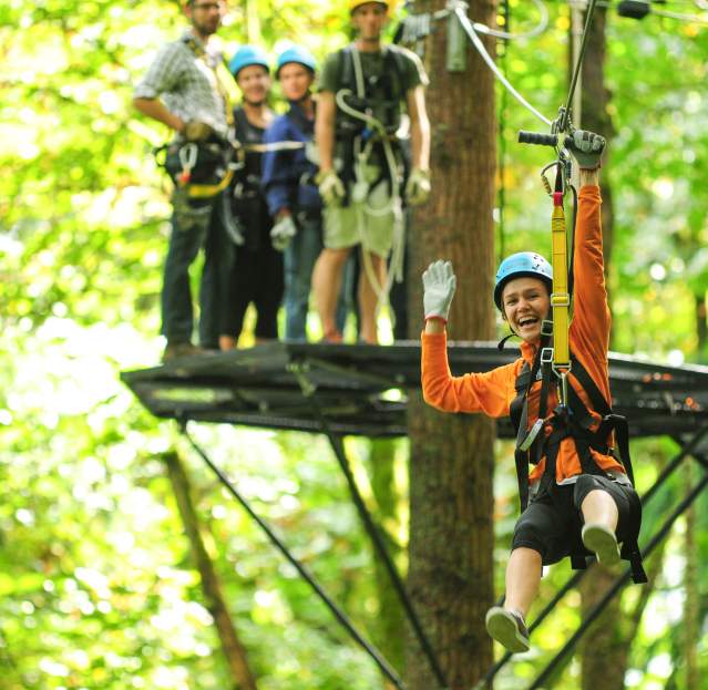 A person in orange zip-line gear is gliding through a lush green forest, with others waiting on a platform in the background.