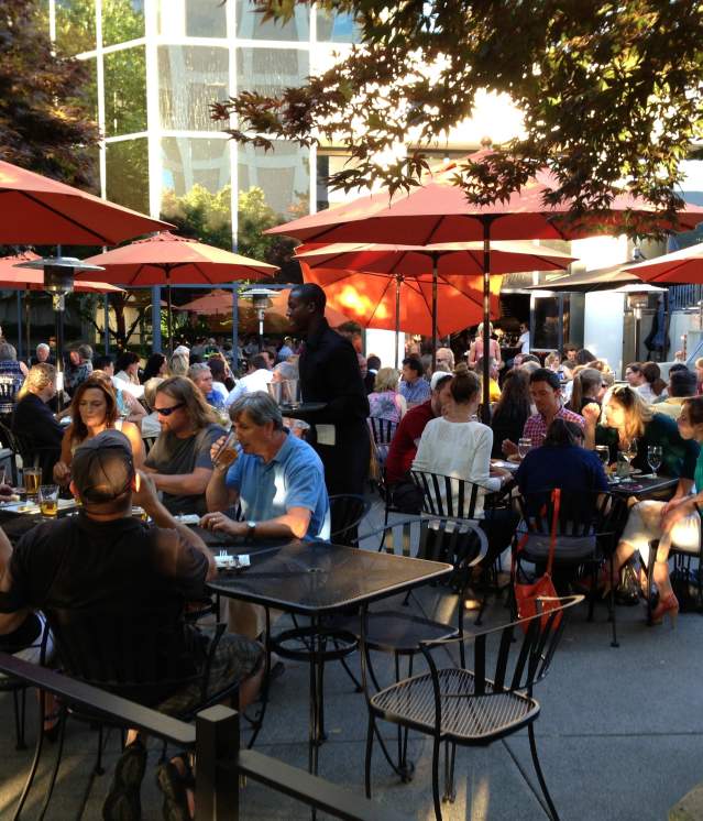 A lively Bellevue outdoor dining area filled with patrons seated under orange umbrellas, enjoying food and drinks in a relaxed atmosphere.