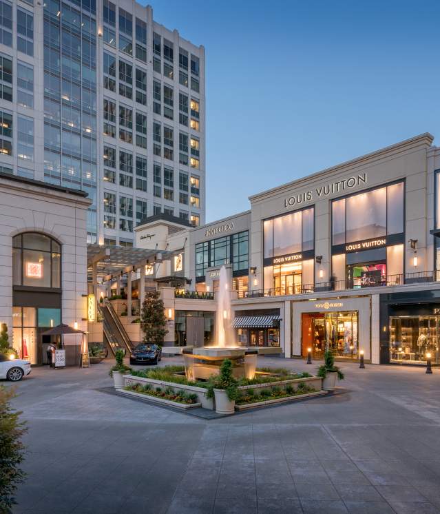 Bravern Bellevue luxury shopping plaza featuring Hermes, Louis Vuitton, Prada, with a fountain and modern architecture at dusk.