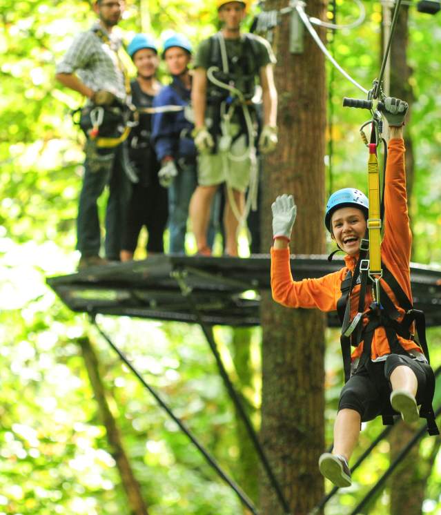 A person in orange zip-line gear is gliding through a lush green forest, with others waiting on a platform in the background.