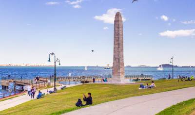 Families sitting on grass on Castle Island near obelisk statue and overlooking Boston Harbor