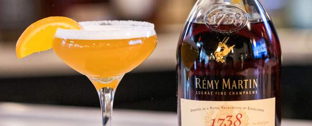 Cocktail glass with orange drink, orange segment, and bottle of cognac on a bar counter