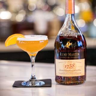 Cocktail glass with orange drink, orange segment, and bottle of cognac on a bar counter