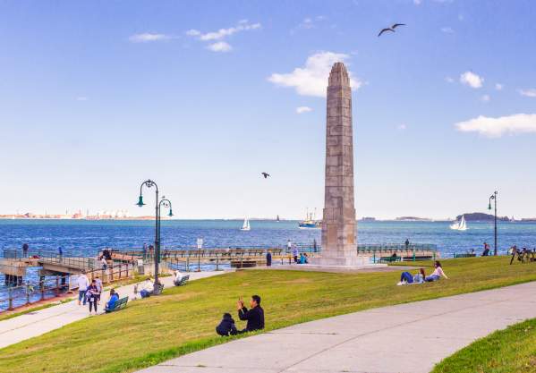 Families sitting on grass on Castle Island near obelisk statue and overlooking Boston Harbor