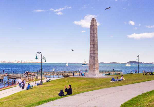 Families sitting on grass on Castle Island near obelisk statue and overlooking Boston Harbor