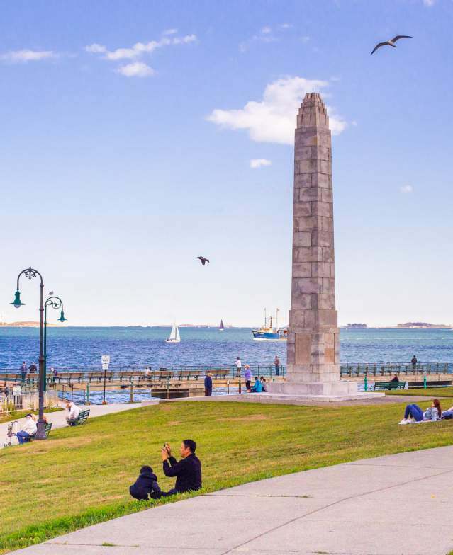 Families sitting on grass on Castle Island near obelisk statue and overlooking Boston Harbor