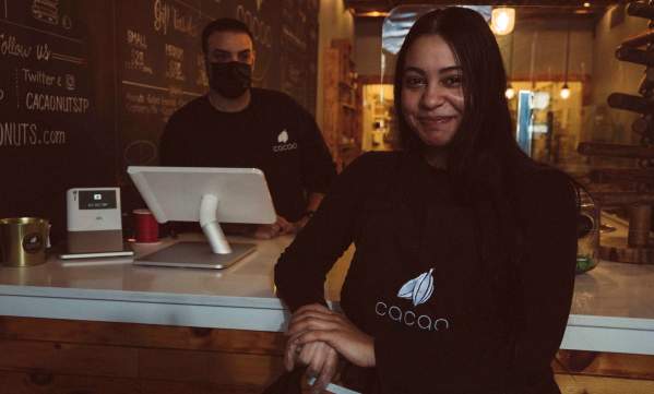 Woman leaning on counter in front of man wearing a cloth mask working behind a register at Cacao