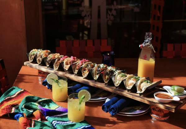 Long row of tacos on a board with two citrusy-looking drinks at Rosa Mexicano