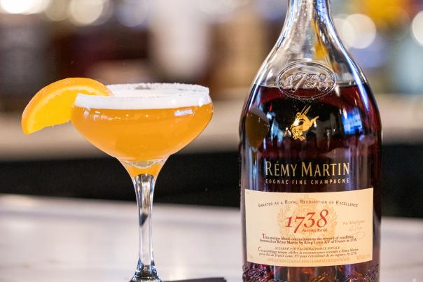 Cocktail glass with orange drink, orange segment, and bottle of cognac on a bar counter