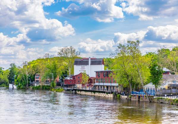 Scenic riverside view at New Hope Riverfront featuring Bucks County Playhouse and colorful buildings amid lush greenery under a partly cloudy sky.