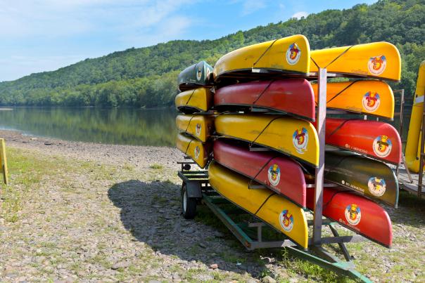 Red and yellow canoes stacked on a canoe trailer with a river and clear blue sky in the background.
