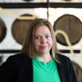 Woman is looking at the camera, wearing a green shirt and black jacket. She is standing in front of barrels and smiling.