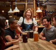 A woman in traditional German dress serving four large beers to a group at Hofbrauhaus Newport