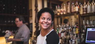 Young Black woman smiling with the bourbon collection of Tousey House Tavern behind her