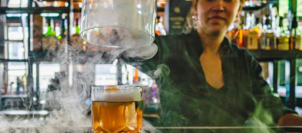 A bartender lifts the dome off a cocktail while smoke swirls around the bar.