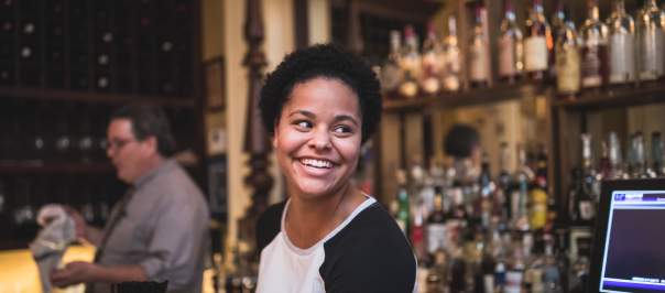 Young Black woman smiling with the bourbon collection of Tousey House Tavern behind her