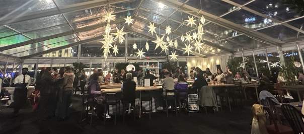 People are gathered in a tent with clear walls and roof. Glowing snowflakes hang from the ceiling.