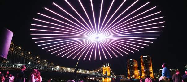 People gather to view a drone light show with the Roebling Suspension Bridge and buildings in the background.