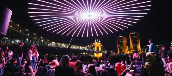 People gather to view a drone light show with the Roebling Suspension Bridge and buildings in the background.