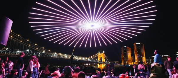 People gather to view a drone light show with the Roebling Suspension Bridge and buildings in the background.