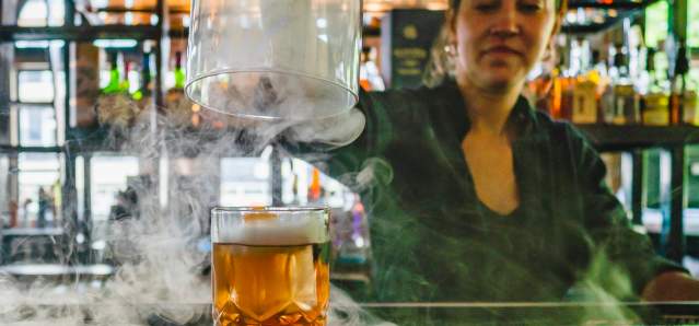 A bartender lifts the dome off a cocktail while smoke swirls around the bar.