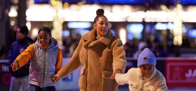 A mom and her two children ice skate while holding hands.