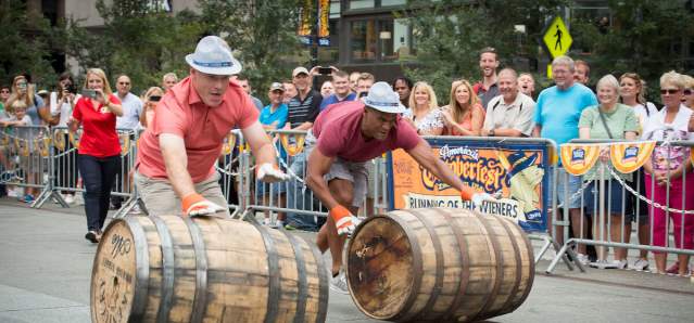 Two people in hats race down a street pushing barrels, while a crowd watches from behind a barricade in the background at Oktoberfest Zinzinnati.