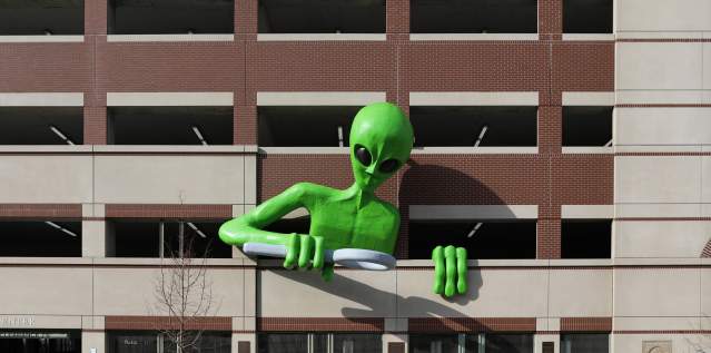 A green alien leans out of a parking garage, looking over the sidewalk with a magnifying glass.
