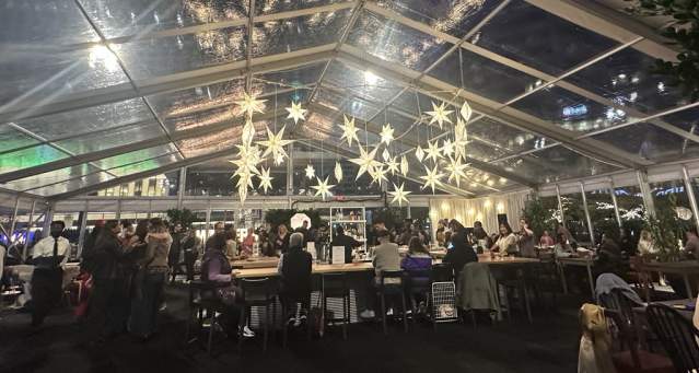 People are gathered in a tent with clear walls and roof. Glowing snowflakes hang from the ceiling.