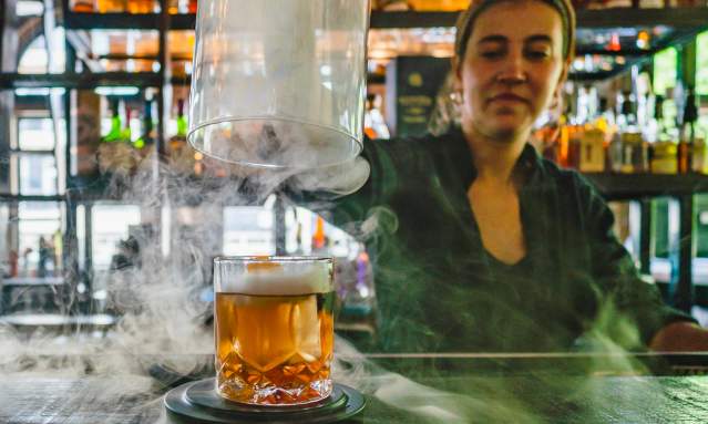 A bartender lifts the dome off a cocktail while smoke swirls around the bar.