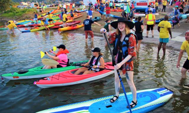 A woman in a hat and life vest stands on a paddleboard in the foreground, while kayaks and a crowd of people are visible in the water and on the shore in the background.