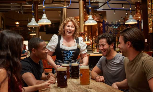 A woman in traditional German dress serving four large beers to a group at Hofbrauhaus Newport