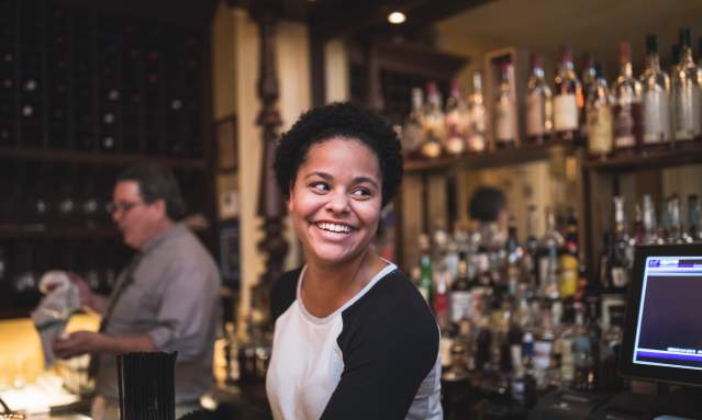 Young Black woman smiling with the bourbon collection of Tousey House Tavern behind her